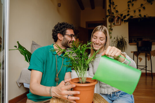 A woman pours water from a watering can into a potted plant held by a man and they laugh while sitting on the floor - Powered by Adobe