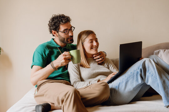 Man holding cup and hugging laughing woman while they sit on bed and look at laptop