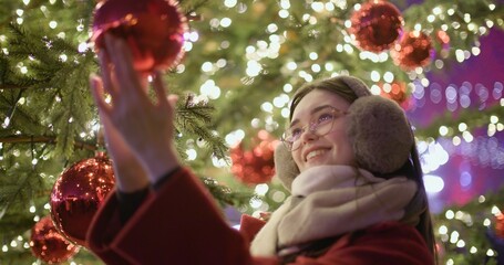 A young beautiful girl in a red coat stands near a Christmas tree with garlands and illuminations in the evening at the European Christmas Market and touches the Christmas tree decorations.
