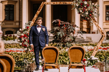 Groom in formal suit stands at floral wedding arch surrounded by vibrant garden flowers, preparing for ceremony with elegant chairs arranged for guests in outdoor setting