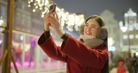 A young beautiful girl in a red coat walks in the evening at the European Christmas Market and talks on the phone against the backdrop of an ice rink with illumination and garlands.
