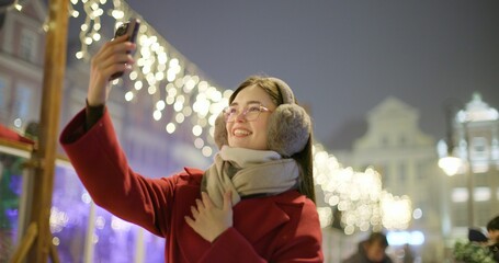 A young beautiful girl in a red coat walks in the evening at the European Christmas Market and talks on the phone against the backdrop of an ice rink with illumination and garlands.