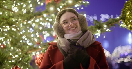 A young girl in a red coat walks in the evening at the European Christmas fair and admires her surroundings while standing against the backdrop of a Christmas tree with garlands and illuminations.