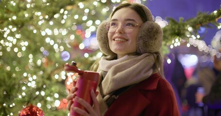 A young beautiful girl in a red coat walks in the evening at the European Christmas Market and holds a thermos cup, standing against the backdrop of a Christmas tree with garlands and illuminations.