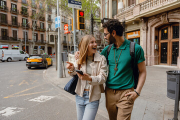 A man hugs a woman who is laughing and pointing in front of him while holding a phone and they...