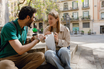 Woman smiling and listening to man holding cup and talking while they sit on stairs and hold package