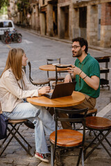 A man is talking to a woman sitting across from him at a table with a laptop on it