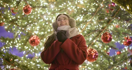 A young girl in a red coat walks in the evening at the European Christmas fair and admires her surroundings while standing against the backdrop of a Christmas tree with garlands and illuminations.