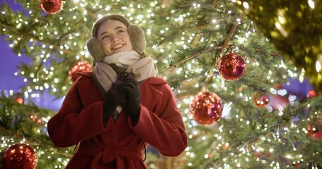 A young girl in a red coat walks in the evening at the European Christmas fair and admires her surroundings while standing against the backdrop of a Christmas tree with garlands and illuminations.