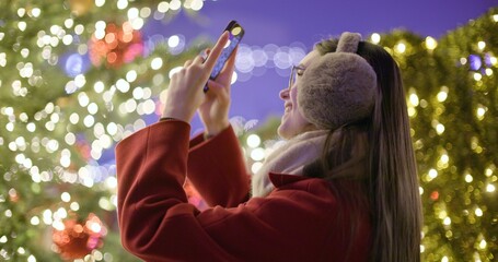 A young beautiful girl in a red coat walks in the evening at the European Christmas Market and takes pictures on a phone while standing against Christmas tree with garlands and illuminations.