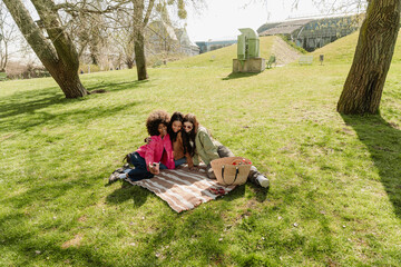 A group of three female friends laughing and sitting on a blanket while posing in front of a phone held by one of them while the other hugs her