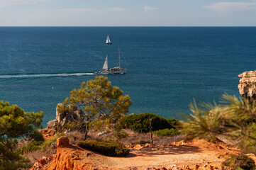 Algarve shoreline with sailboat and excursion yacht on the Atlantic, seen from cliff path. Perfect for tourism platforms, destination guides, booking websites.