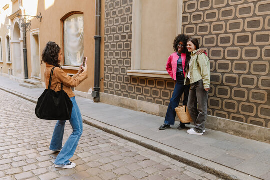 A woman stands and takes a photo on her phone of two female friends smiling and posing near a wall while one of them hugs the other