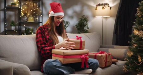A woman wearing a Santa hat is opening Christmas gifts on a sofa in a cozy living room. A person holds wrapped gifts near a decorated Christmas tree, creating a festive holiday scene