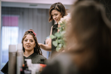 Hispanic woman in salon chair smiling while stylist works on her hair, surrounded by beauty products and tools, creating a vibrant atmosphere of self-care and pampering