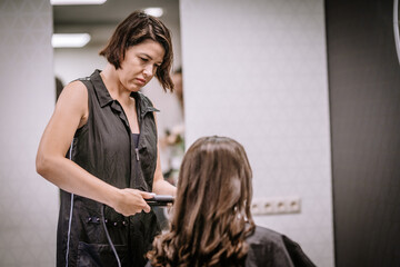Female hairstylist with dark hair is styling long hair of client in modern salon, showcasing professional techniques and contemporary beauty environment