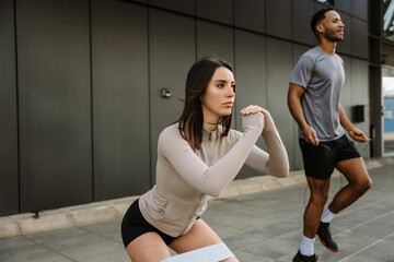 A female athlete looks ahead and squats with elastic bands on her thighs while a male athlete jumps rope next to her