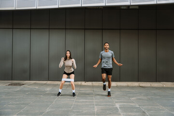 A female athlete squats with a rubber band on her legs while a male athlete jumps rope next to her