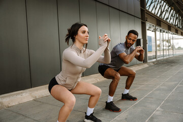 Naklejka premium A male athlete talks and looks at a female athlete who looks ahead while they squat and hold their hands together in front of them