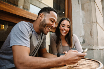 Fototapeta premium Male and female athletes laughing and sitting at a table while holding cups and looking at the phone he is showing