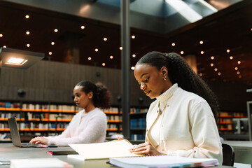 Two female students sit at a table while one reads the cover of a folder and the other types on a laptop