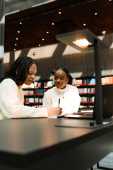 A female student is talking to a female student who is writing in a notebook and sitting next to her at a table