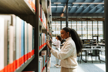 Two female students are looking for books on bookshelves they are standing next to while one of them is holding a book