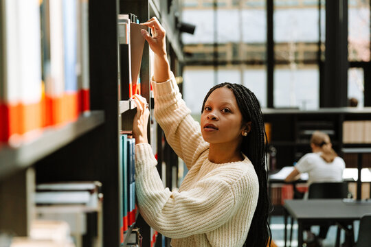 Female student standing and taking out a book while leaning on a bookshelf and looking away - Powered by Adobe