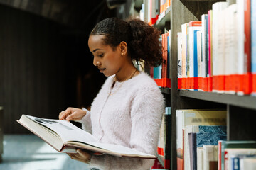 Female student holding a book and standing near bookshelves while reading