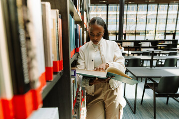 Female student leaning on bookshelves and holding a book she is reading while pointing with her finger