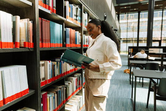 Female student smiling and reading a book she is holding while standing near bookshelves