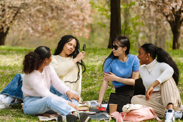 A female student looks into a magnifying glass while sitting on a blanket next to a group of three female students, one of whom is laughing
