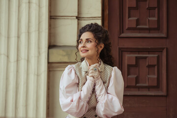 Fototapeta premium An elegant woman in a vintage dress sits next to an old stone column with a very happy expression on her face.