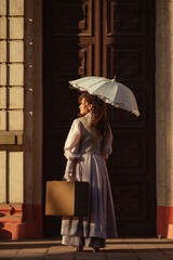 Fototapeta premium A woman in an antique dress with an umbrella and a suitcase stands in front of the doors of an old cathedral.