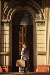 Fototapeta premium A woman in a vintage dress with a suitcase stands by ancient columns at dusk.