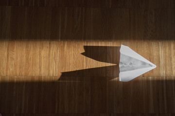 Paper airplane casting a distinct shadow across a sunlit wooden floor captured from above in mid-afternoon light