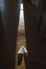 A solitary paper airplane casting shadows on the wooden floor, illuminated by a narrow beam of sunlight filtering through an open doorway