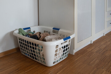 A cluttered laundry basket filled with assorted clothing, positioned neatly against a wall in a modern interior with wooden flooring and a beige wardrobe