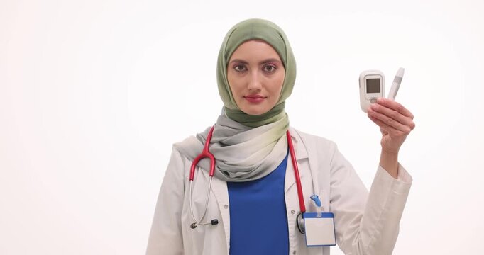 Muslim doctor holds glucose monitoring device and glass with sugar cubes on white background. Physician in hijab explains dietary risks. Slow motion
