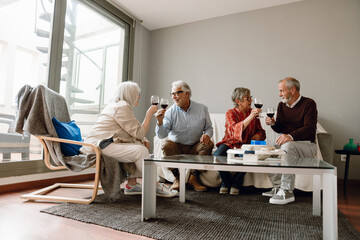 A group of four friends clink glasses and talk in pairs while one of them sits on a chair and three of them sit on a sofa