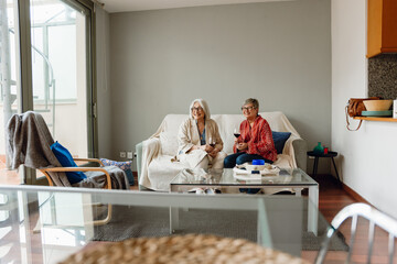 Two female friends laughing and looking away while sitting on a sofa and holding glasses