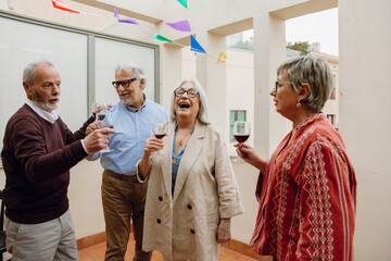 A group of four friends stand and hold glasses while talking and laughing and one of them puts his...
