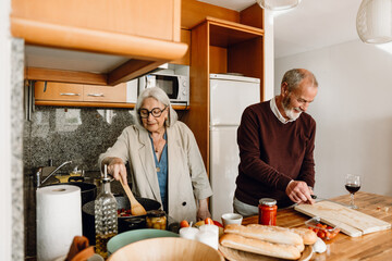 A woman stirs a frying pan with a spatula while a man stands next to her at the table and smiles, holding a knife and cutting board