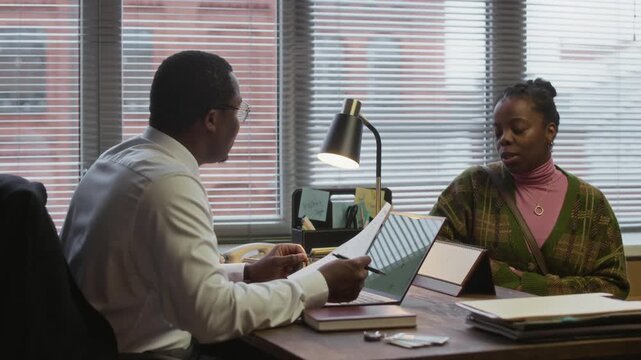 Medium shot of young black parent talking to school principal holing report card while sitting at desk in modern office