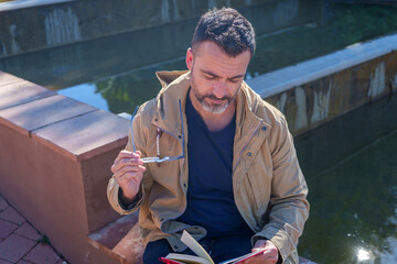 Mature man reading a book outdoors, holding eyeglasses, finding a quiet moment for reflection and learning