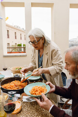 A woman stands and holds a spatula and fork in a frying pan while a man holds a plate and sits at a...