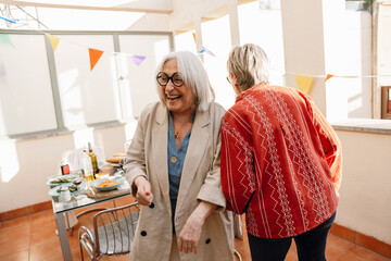 A woman stands and laughs as her female friend walks beside her and they touch shoulders
