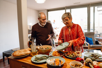 A man holds a bottle and talks to a woman who is smiling and tossing spaghetti from plate to plate while they stand at the table