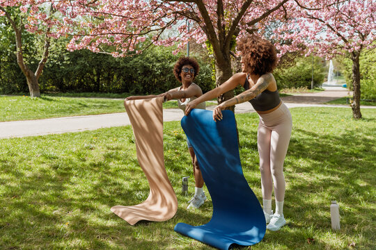 Two female friends spread out mats on the grass while standing and laughing while looking at each other