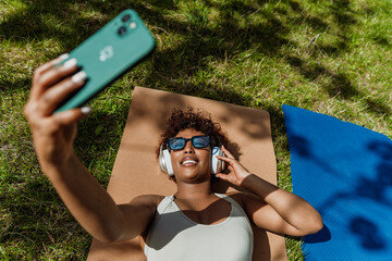 A woman looks at her phone and lies on a mat on the grass while listening to headphones
